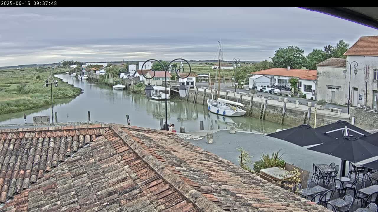Overlooking a calm river in a small town, boats are moored along the waterfront under an overcast sky, with outdoor seating visible at a nearby establishment.