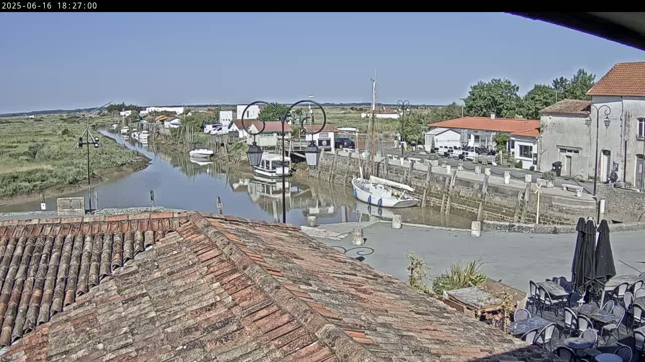 A sunny view overlooking a calm river flowing through a small town with docked boats, buildings, and outdoor seating.