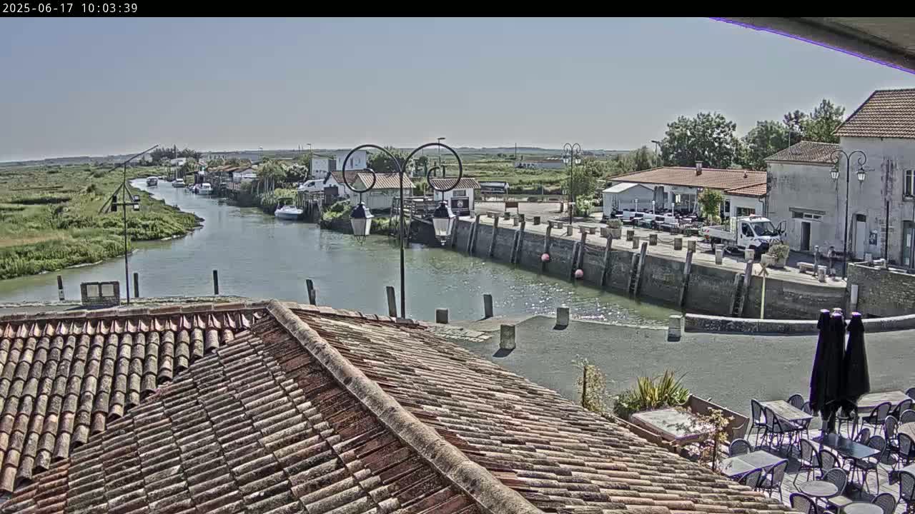 A calm river flows past a waterfront village under a clear sky, where boats are moored and outdoor seating is set up next to a building with a terracotta tile roof.