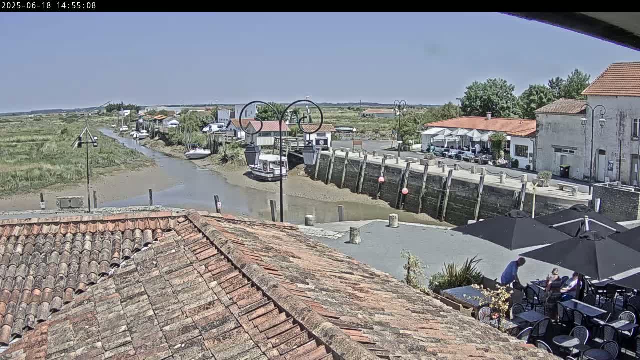 A sunny view overlooking a calm waterway in a small harbor town, with boats moored along the banks and patrons seated at outdoor tables under umbrellas at a nearby restaurant.
