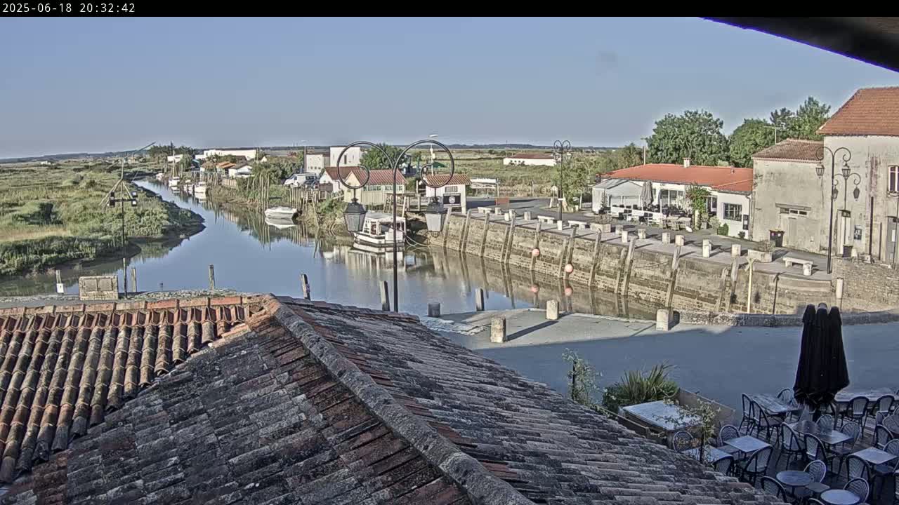 A calm river flows through a quaint village under a clear, sunny sky, past buildings with terracotta roofs and outdoor seating, and a small boat is moored on the water.