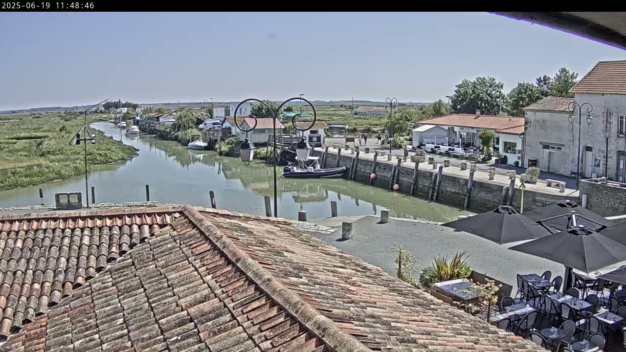 A sunny view of a waterfront village with a calm river, boats docked along a stone quay, buildings with terracotta roofs, and outdoor seating under umbrellas at a restaurant.