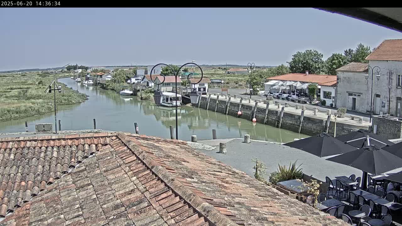 A canal winds through a village under a clear, sunny sky, passing by restaurants with outdoor seating and docked boats.
