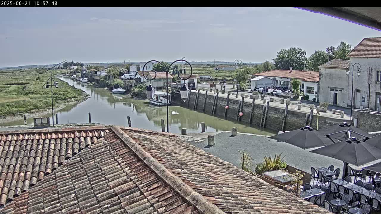 A calm waterway winds through a village on a sunny day, boats are moored along the banks, and outdoor seating is set up at a restaurant near a stone quay.