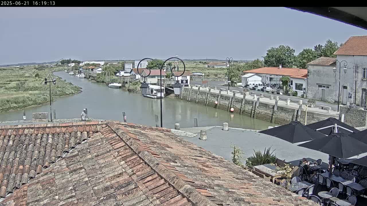 A canal-side village is viewed from a tiled roof on a sunny day, with boats moored along the waterway and outdoor seating at a nearby cafe.