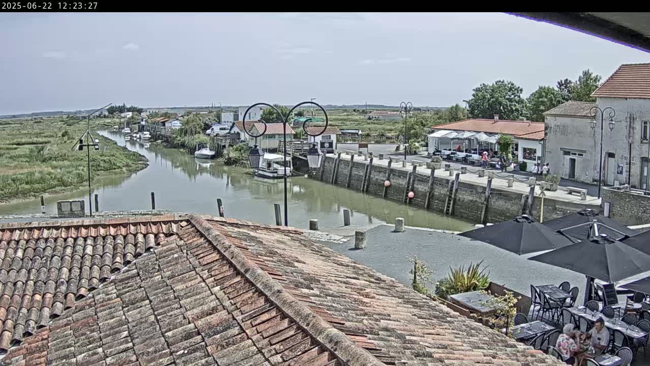 A canal flows through a village on a sunny day, boats are moored along its banks, and outdoor seating areas are set up near waterfront buildings.