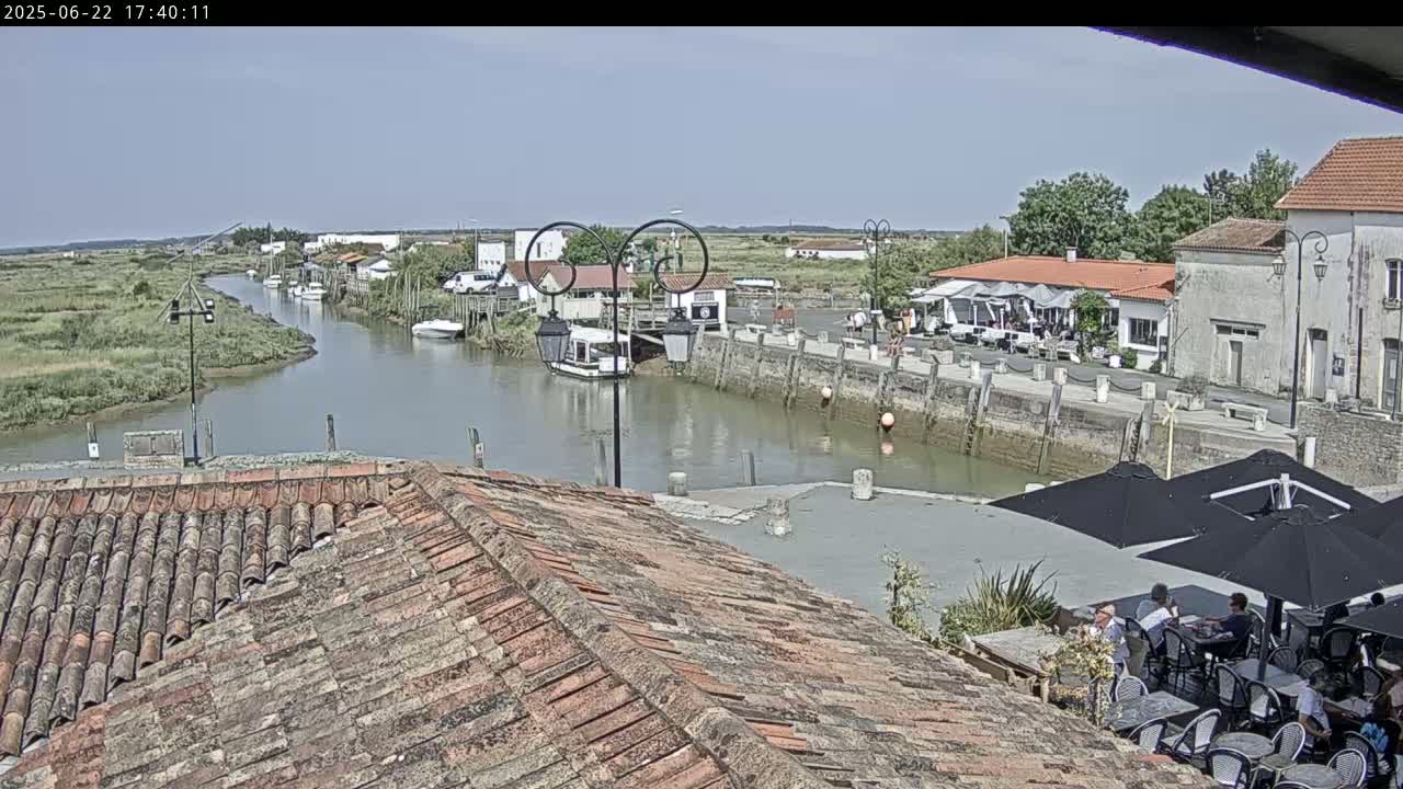 A tranquil waterway winds through a village under a clear sky, where people dine outdoors at a waterfront restaurant.