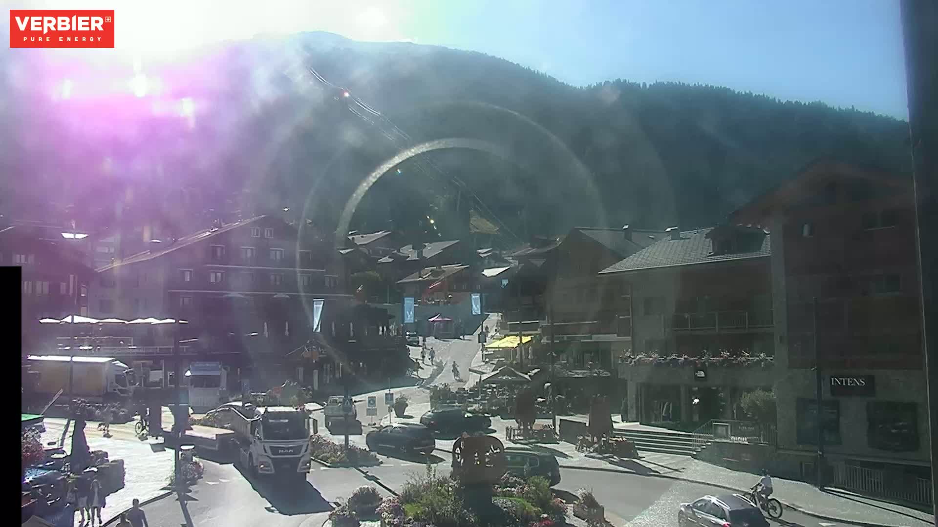 A sunny day in a mountain town shows a street scene with several buildings, vehicles, and pedestrians, with a mountain range in the background.