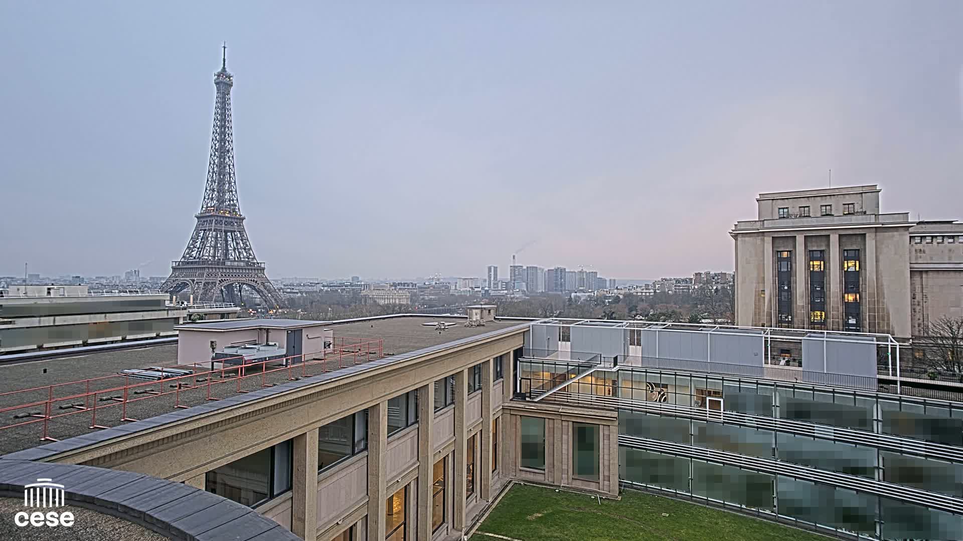 A rooftop view of Parisian buildings, including the Eiffel Tower in the distance, under an overcast sky.