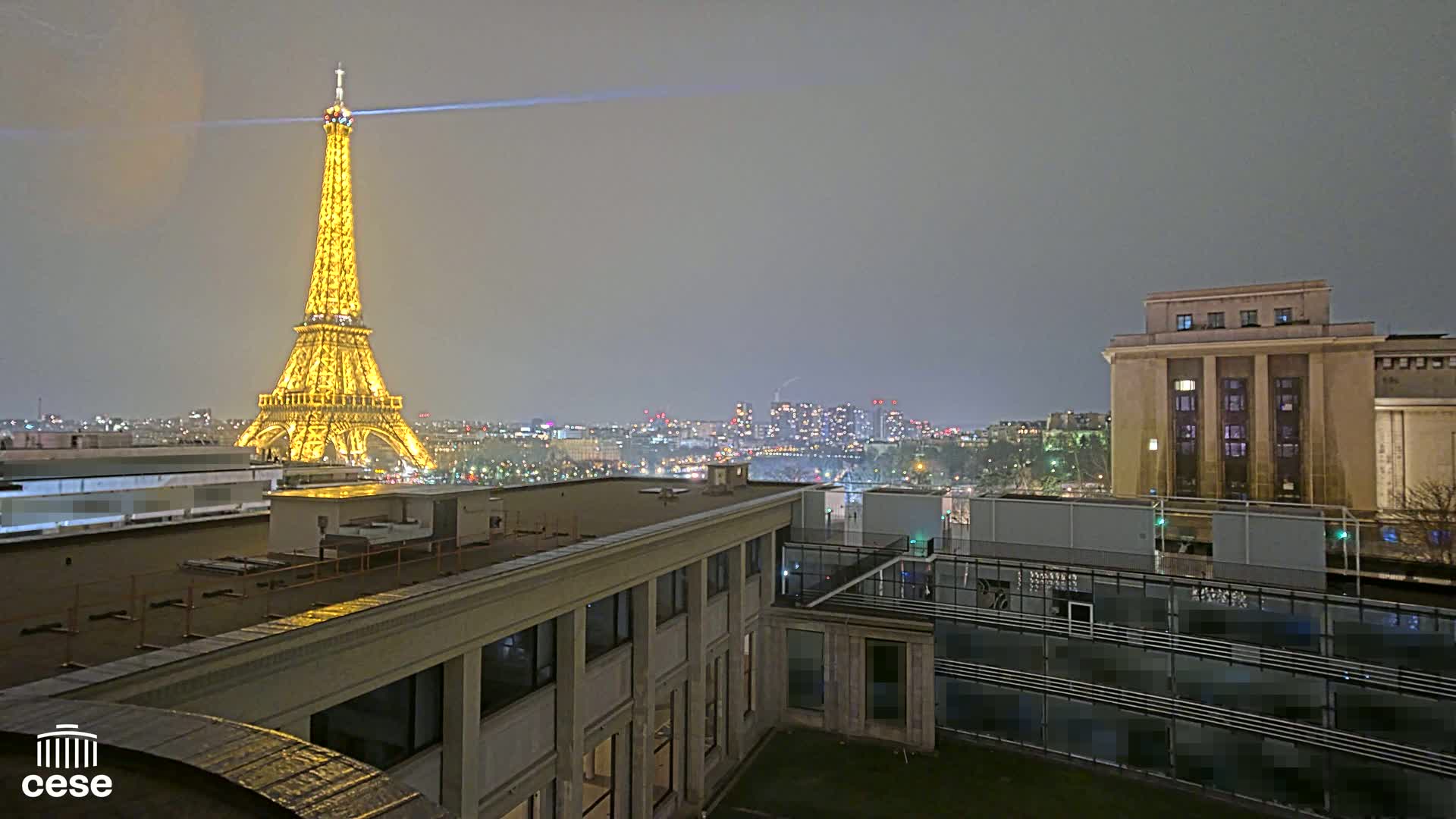 A nighttime view of the Eiffel Tower illuminated, overlooking a city skyline under a cloudy sky.