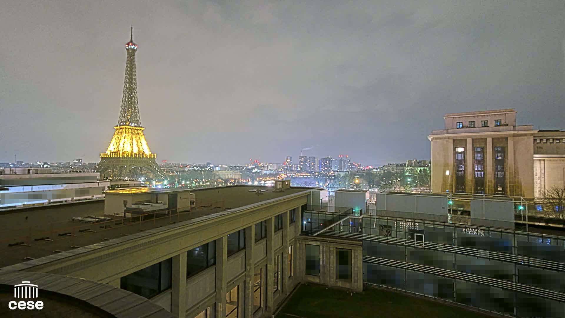 A nighttime cityscape view from a rooftop shows the Eiffel Tower illuminated, city lights in the distance under a cloudy sky, and a large building in the foreground.