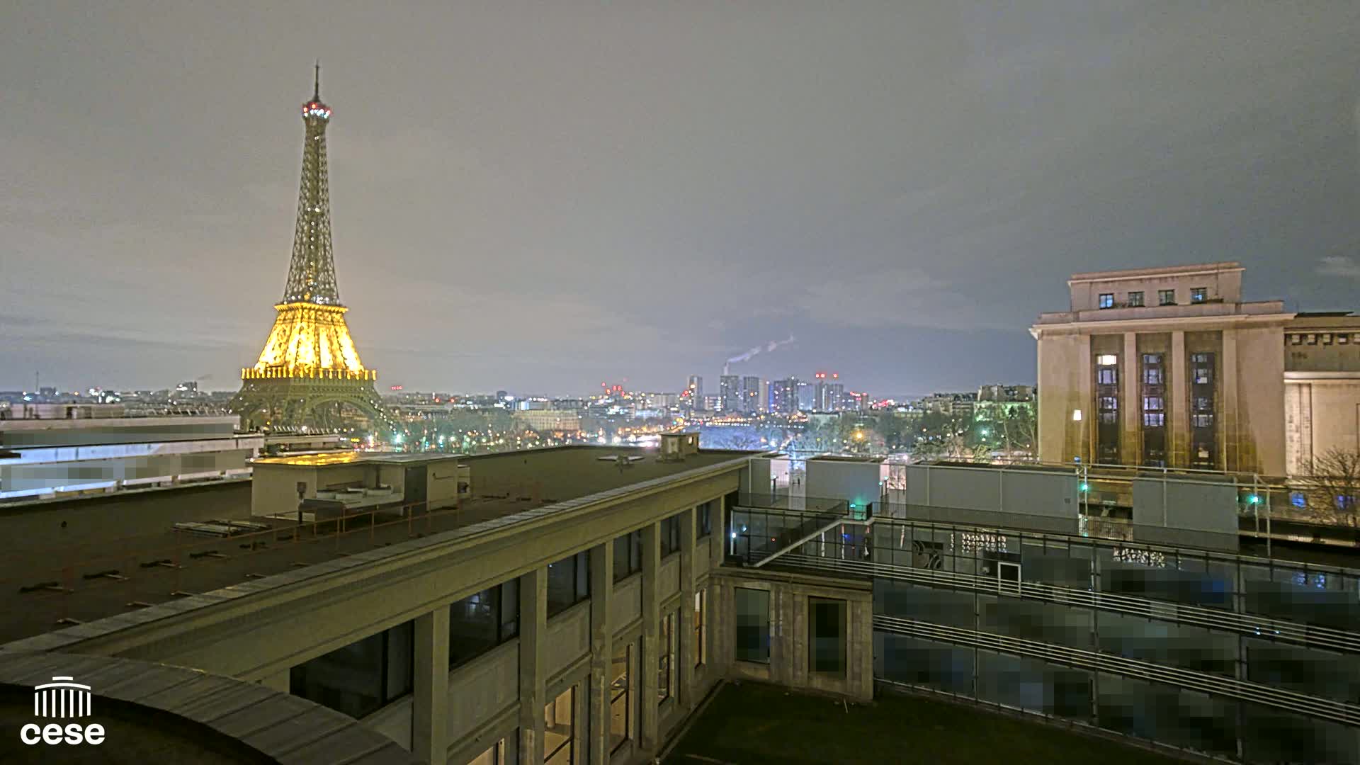 A nighttime view of the Eiffel Tower illuminated, city skyline, and buildings, under a cloudy sky.