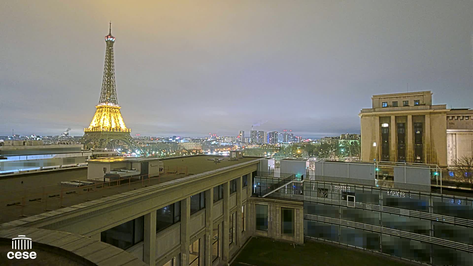 A nighttime view of the Eiffel Tower illuminated, a cityscape, and a large building, under a cloudy sky.