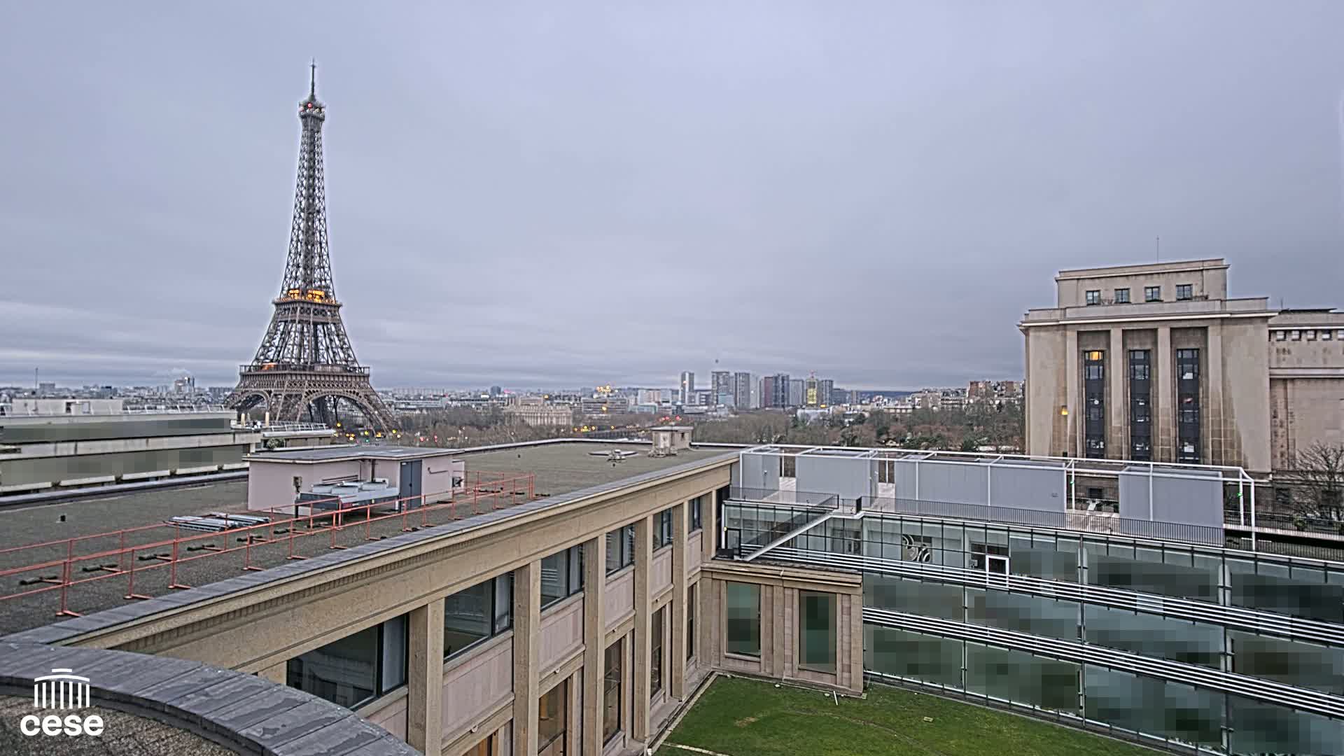 A view from a rooftop shows the Eiffel Tower in the distance under a cloudy sky, with modern and older buildings in the foreground.