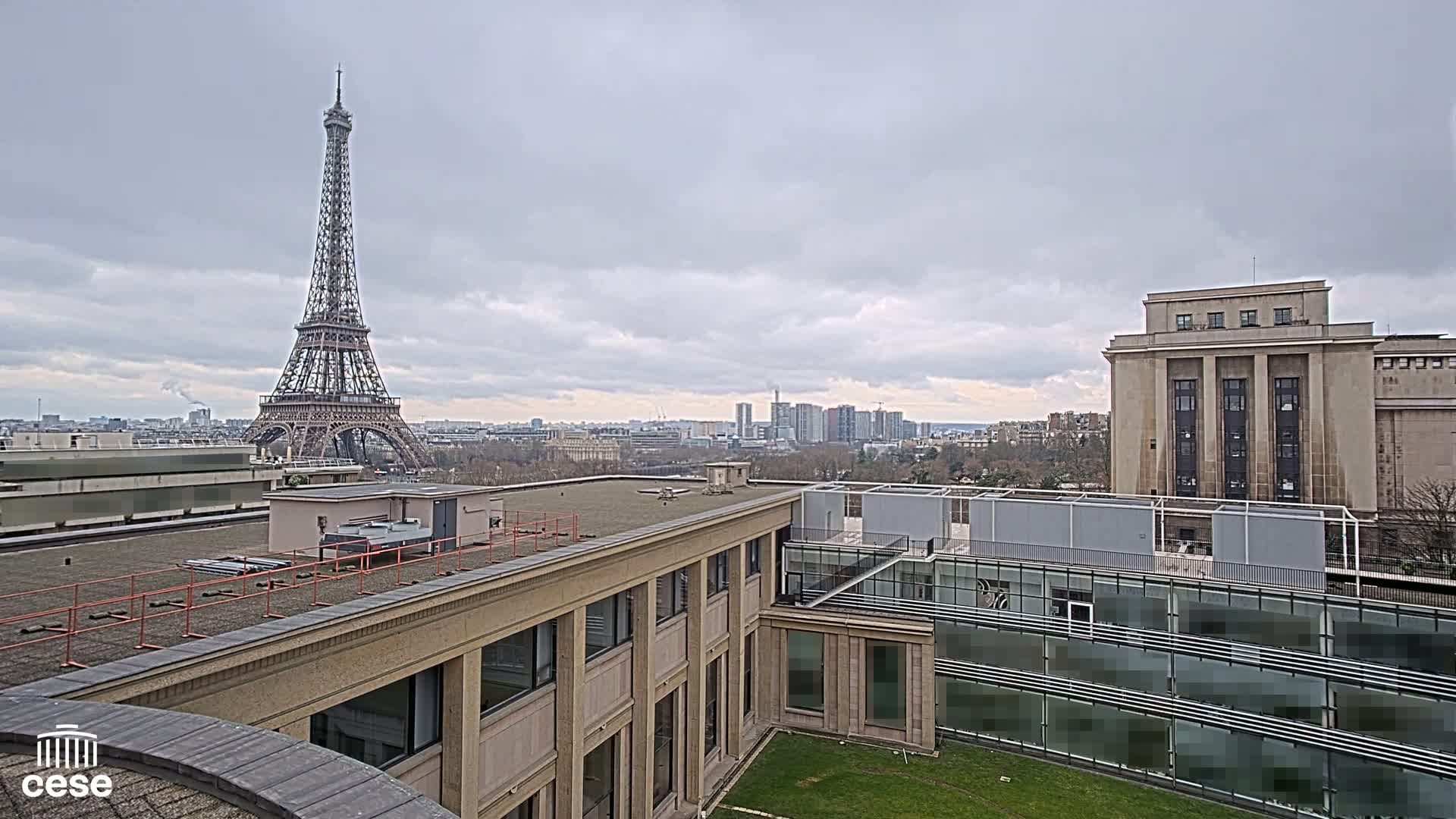 A view from a rooftop shows the Eiffel Tower in the distance under a cloudy sky, with modern and classical buildings in the foreground.