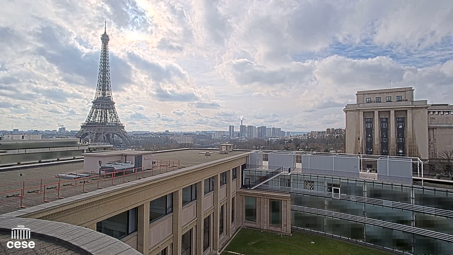 A Parisian cityscape under a partly cloudy sky features the Eiffel Tower in the distance,  seen from atop a building with a flat roof and modern additions.