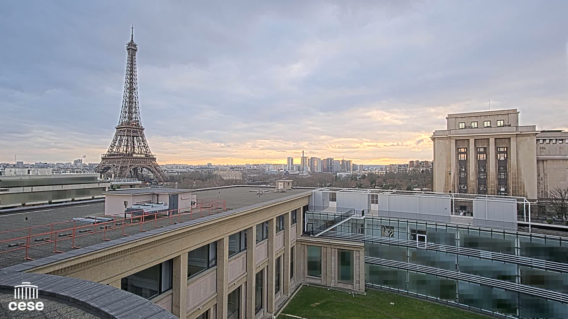 Under a partly cloudy sky, an elevated view shows the Eiffel Tower in the distance and a cityscape with modern and classical buildings.