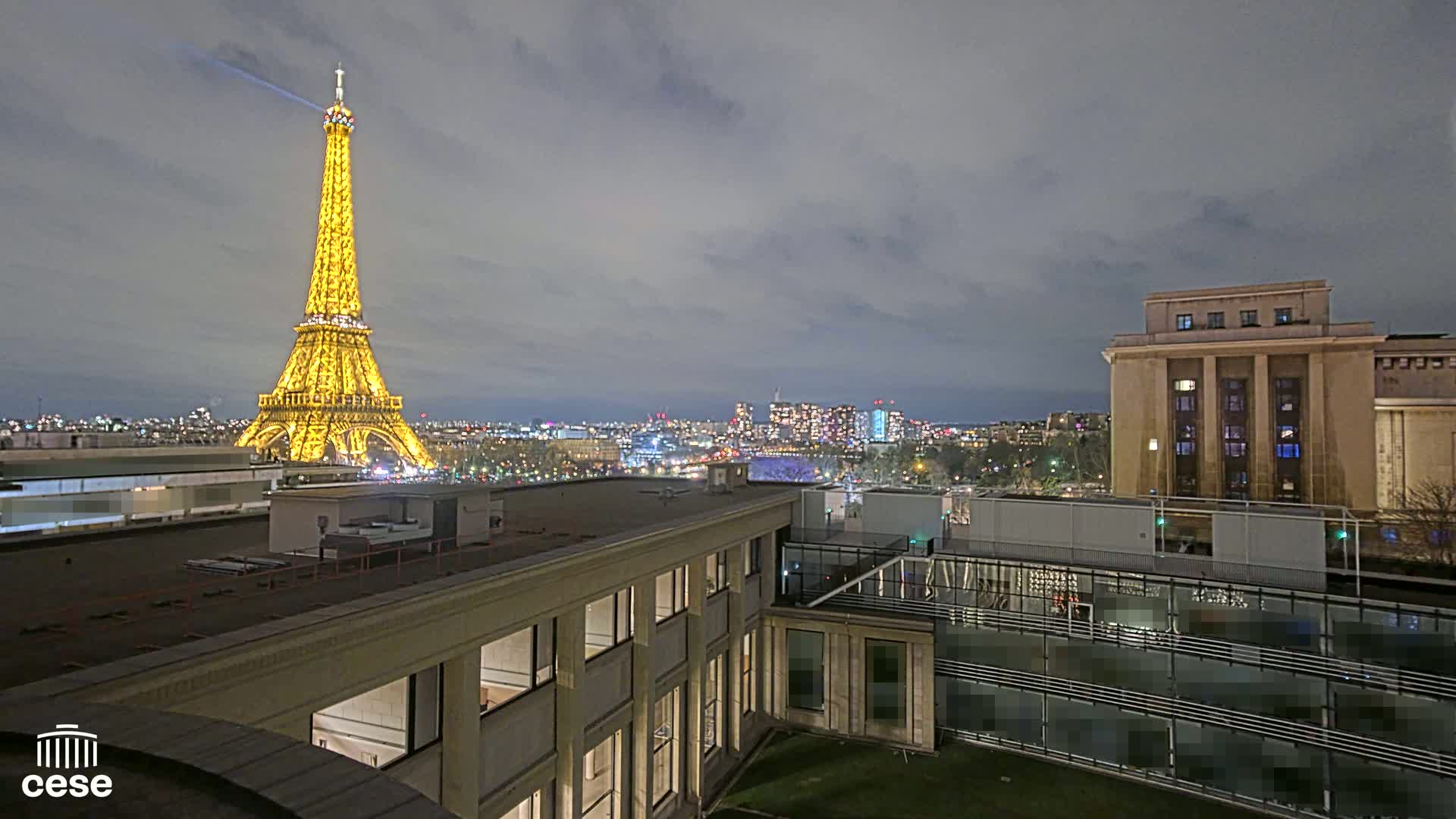 A nighttime view of the Eiffel Tower illuminated in gold, overlooking a city under a cloudy sky.