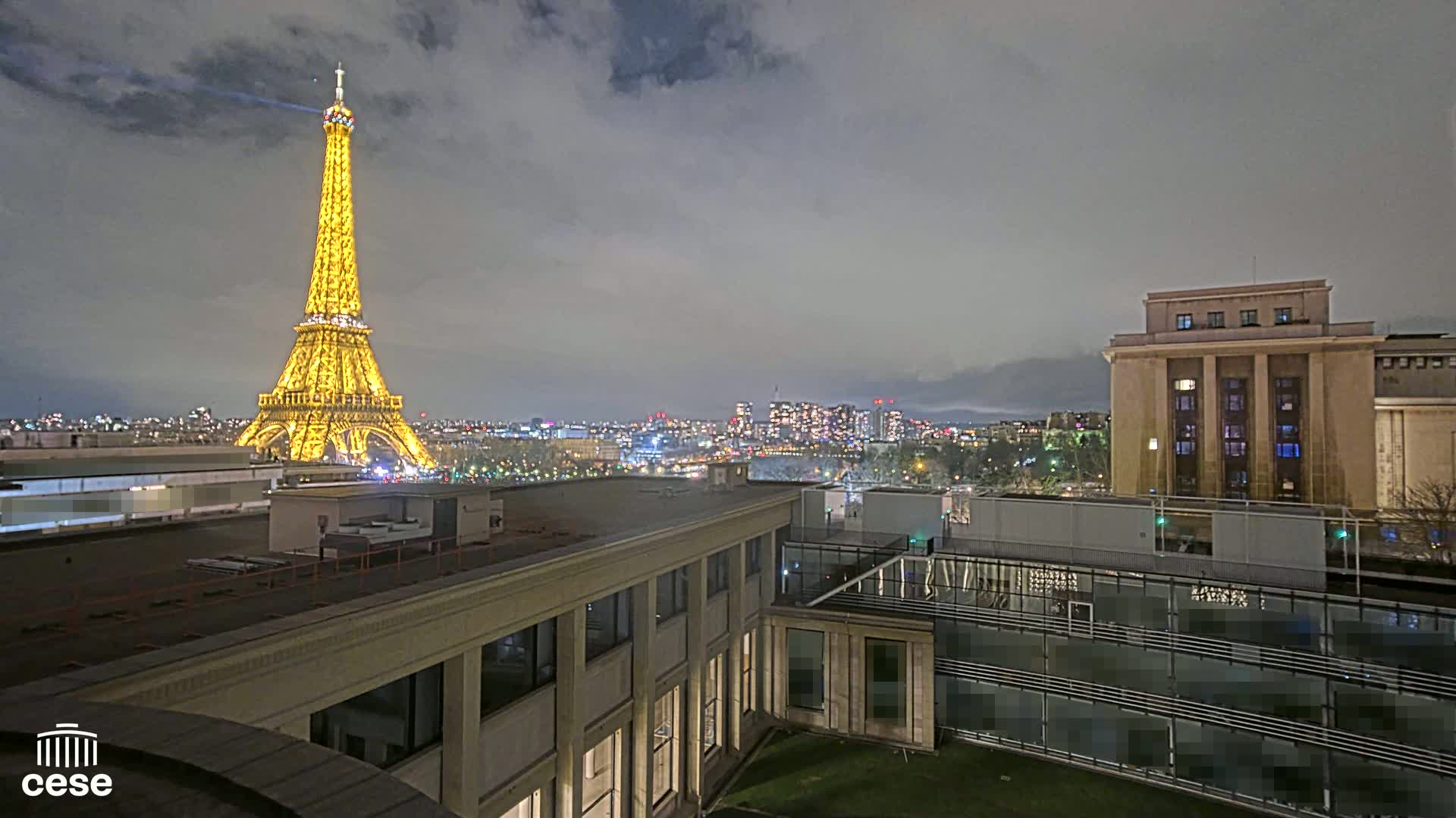 A nighttime view of the Eiffel Tower illuminated, a city skyline, and several buildings under a cloudy sky.
