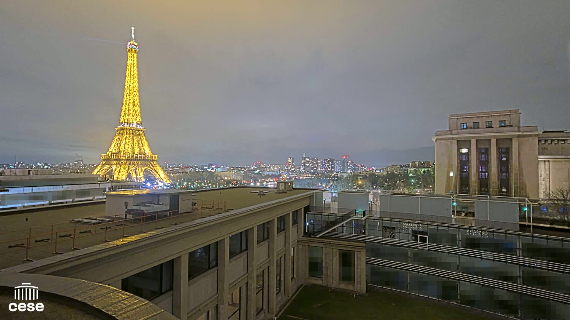 A nighttime view of the Eiffel Tower illuminated, a city skyline, and several buildings under a cloudy sky.