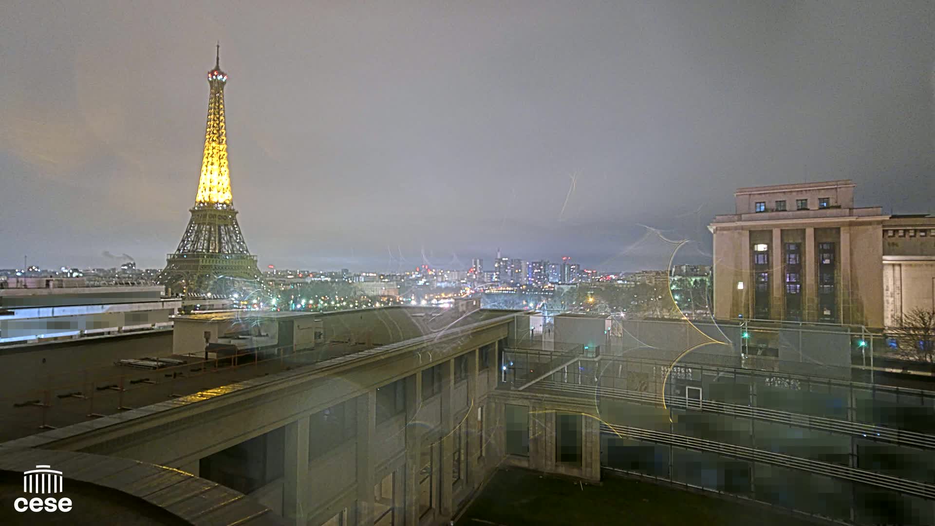 A nighttime view of the Eiffel Tower illuminated, a city skyline, and several buildings under a hazy sky.