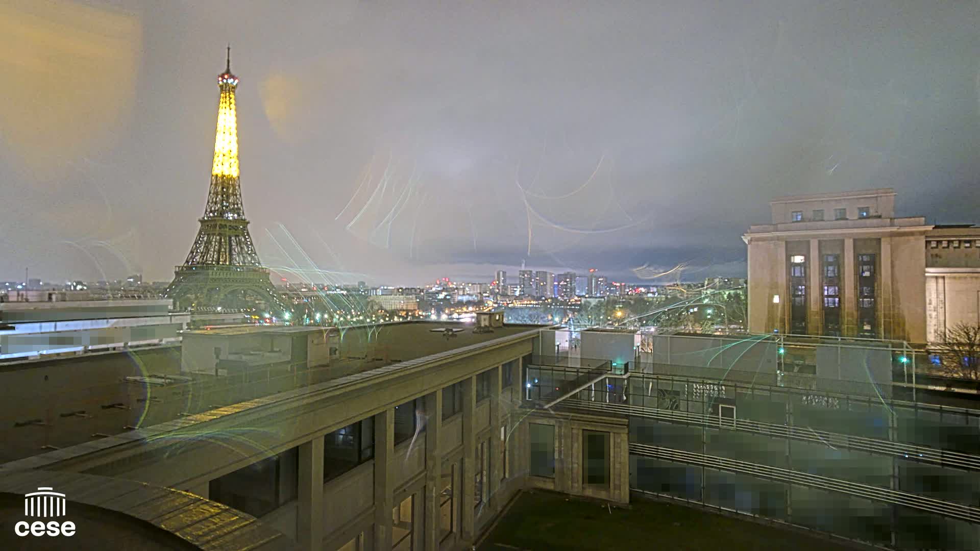 A nighttime cityscape view shows the Eiffel Tower illuminated, a large building, and other city structures under a cloudy sky with light streaks possibly from long-exposure photography.