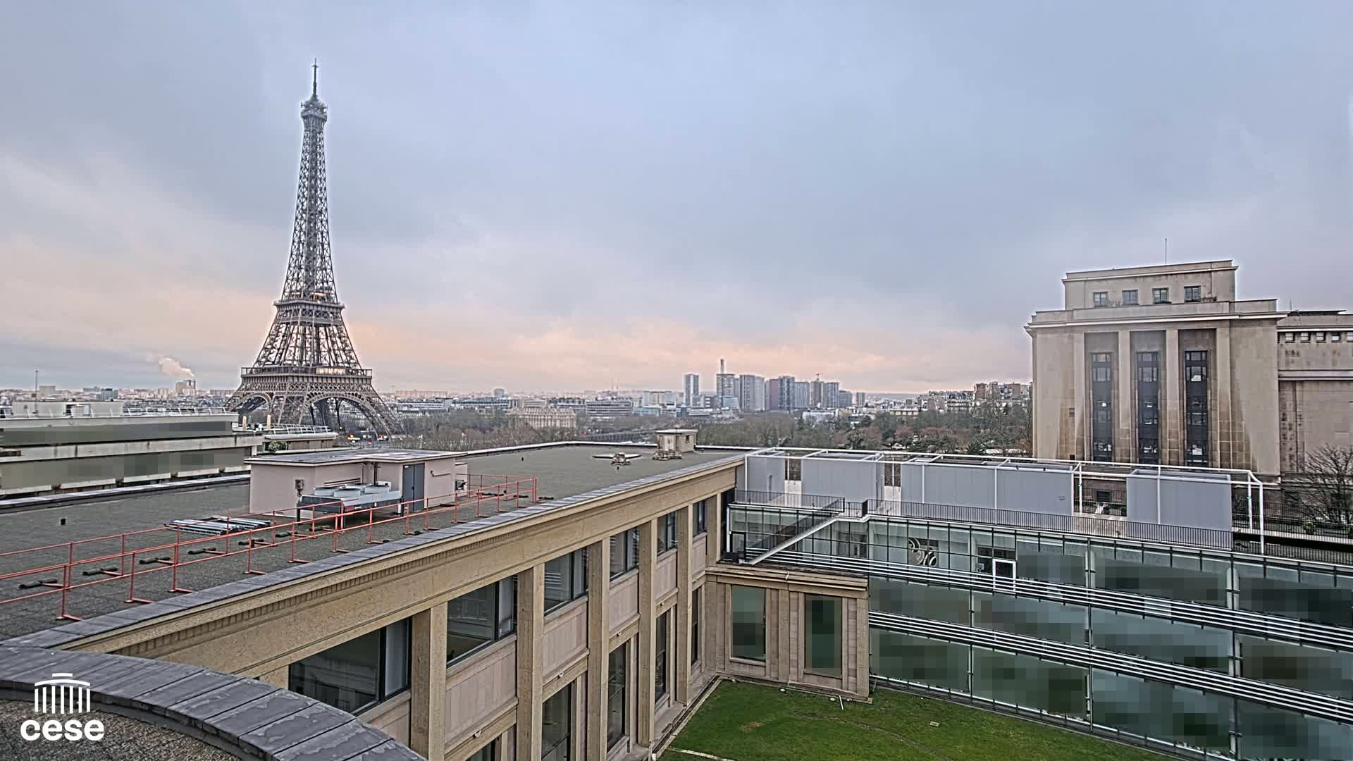 A rooftop view of Parisian buildings, including the Eiffel Tower in the distance, under a cloudy sky.