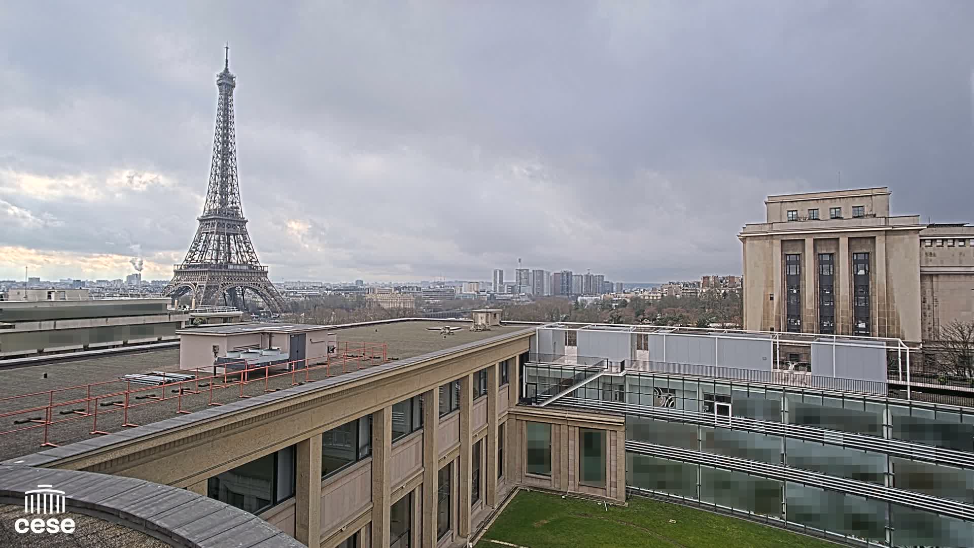 A view from a rooftop shows the Eiffel Tower in the distance under a cloudy sky, with other buildings and a small grassy courtyard in the foreground.
