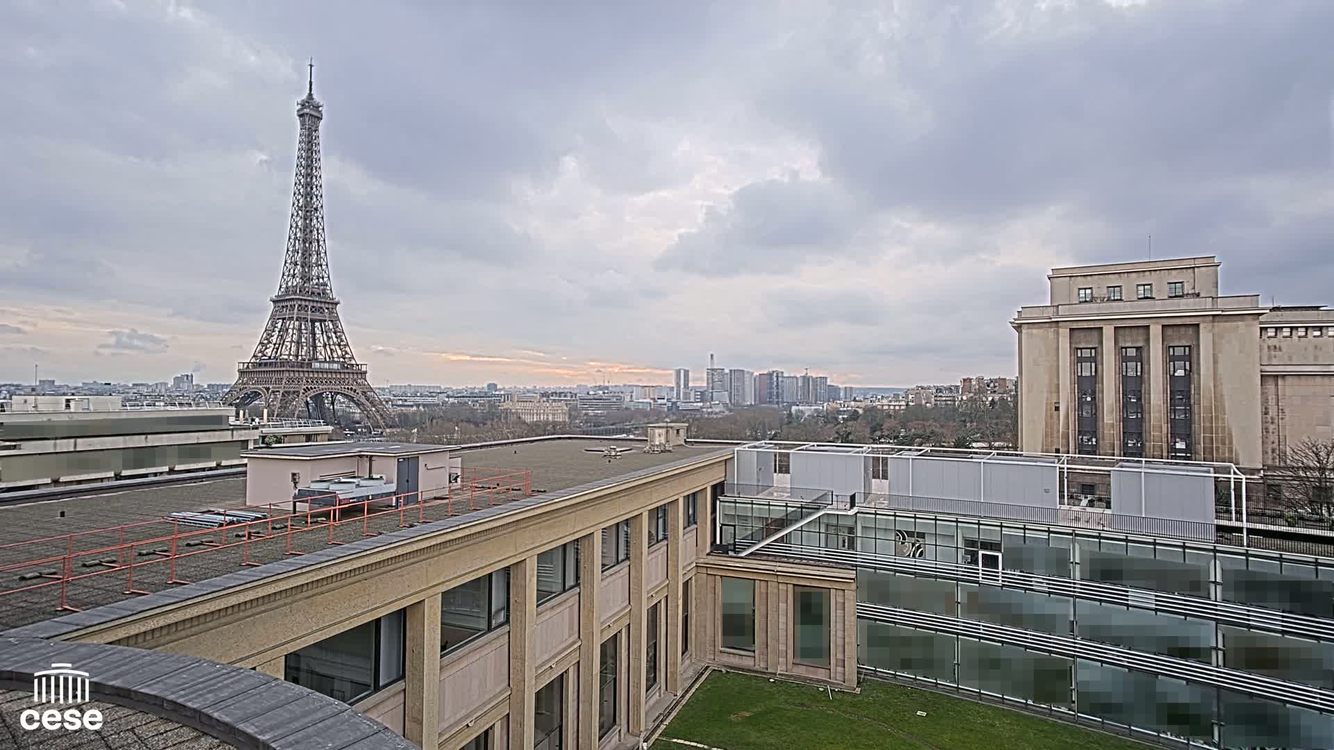 A rooftop view of the Eiffel Tower in the distance under a cloudy sky, with modern and classical buildings in the foreground.