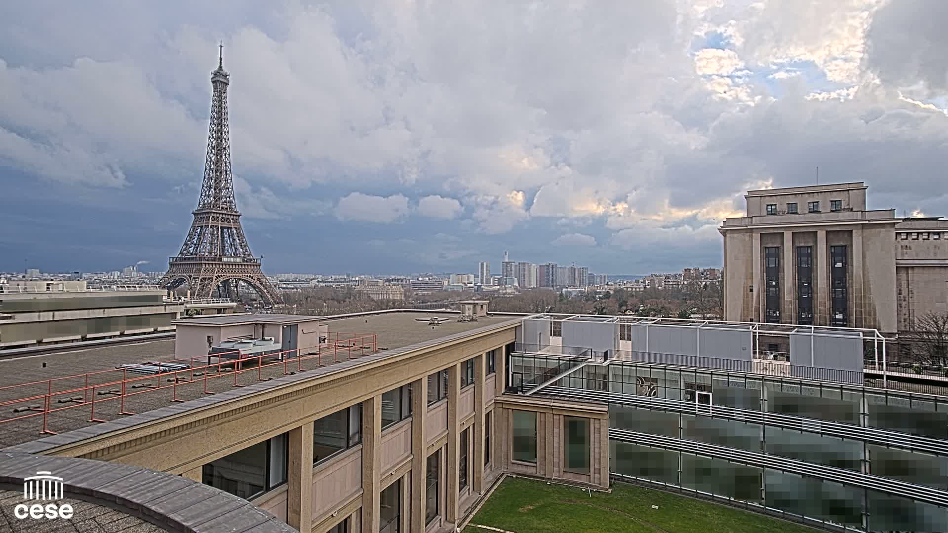 A Parisian rooftop view shows the Eiffel Tower in the distance under a partly cloudy sky, with a modern building and a classical building in the foreground.