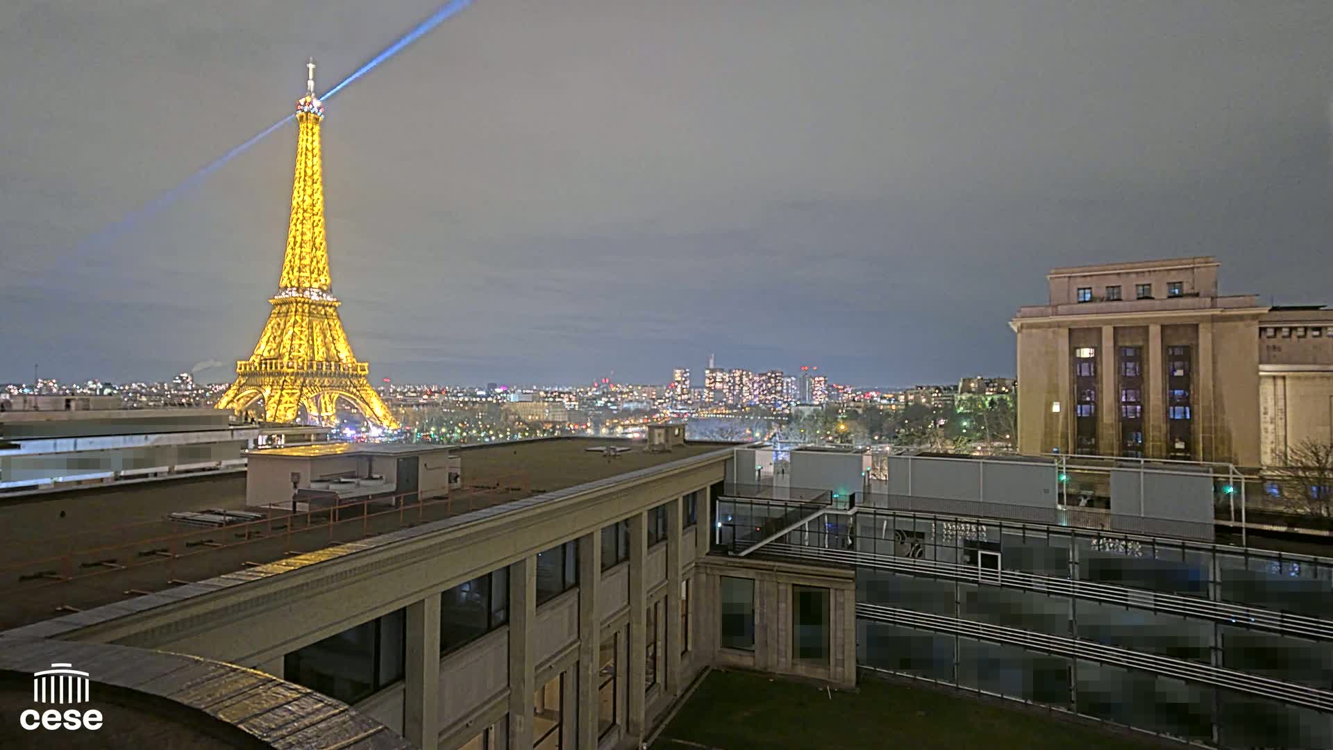 A nighttime view of the Eiffel Tower illuminated,  a city skyline, and rooftops of buildings under a cloudy sky.