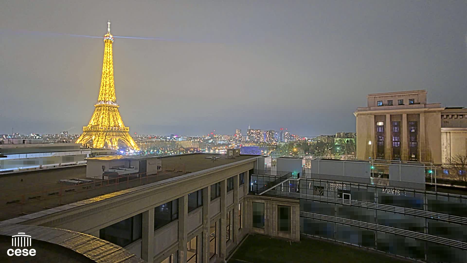 A brightly lit Eiffel Tower is visible in the distance at night, from a rooftop vantage point overlooking a city skyline under a cloudy sky.