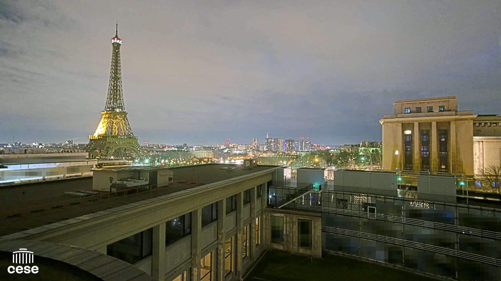 A nighttime view of the Eiffel Tower illuminated against a dusky sky, overlooking a city skyline and buildings from a rooftop vantage point.