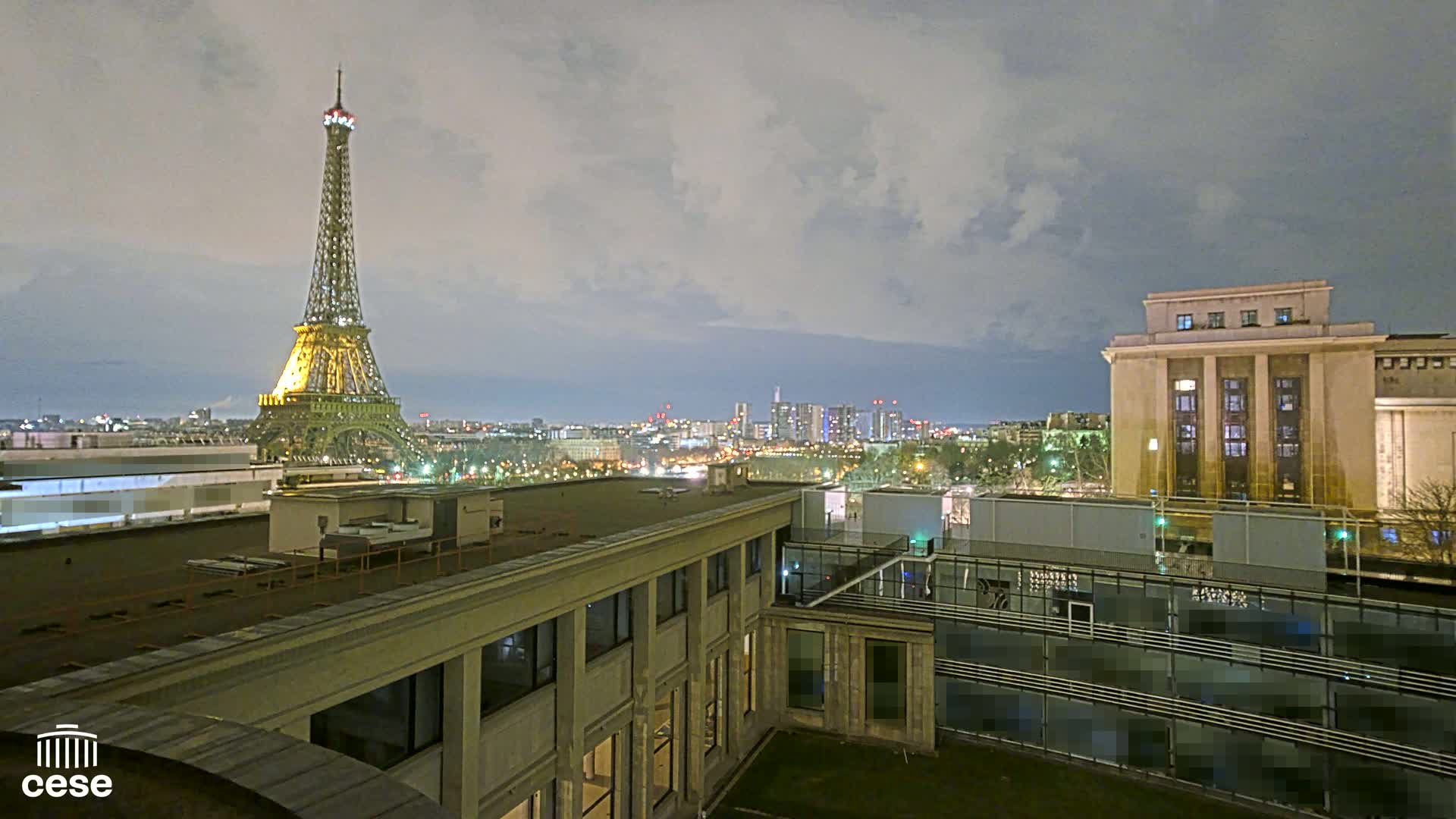 A nighttime view of the Eiffel Tower from a rooftop, under a cloudy sky, shows a cityscape with a large building in the foreground.