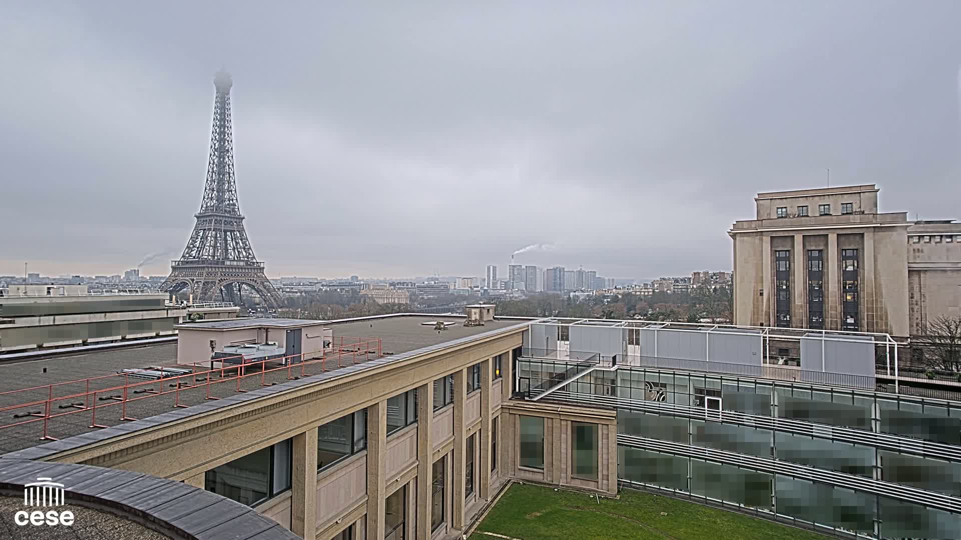 A view from a rooftop shows the Eiffel Tower in the distance under a cloudy sky, with modern and classical buildings in the foreground.