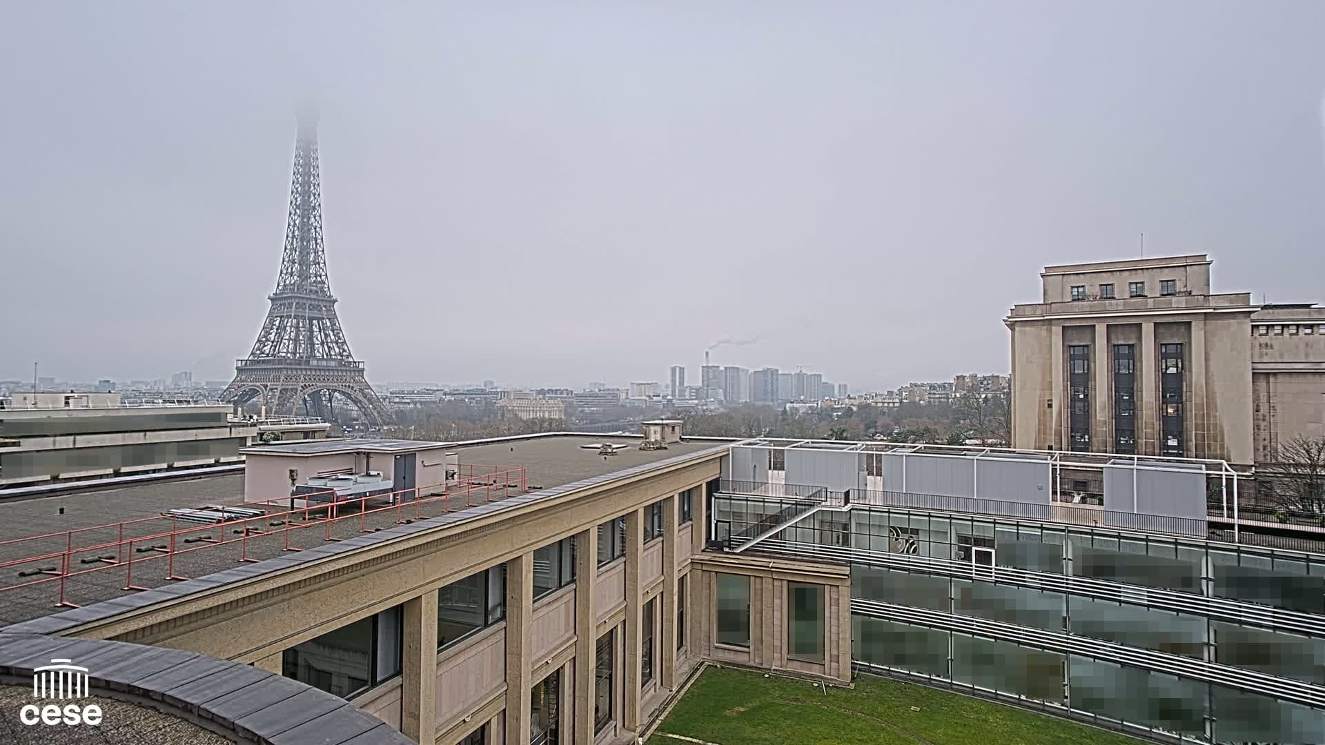 A foggy cityscape view shows the Eiffel Tower in the distance, alongside modern and classical buildings, with a grassy courtyard in the foreground.