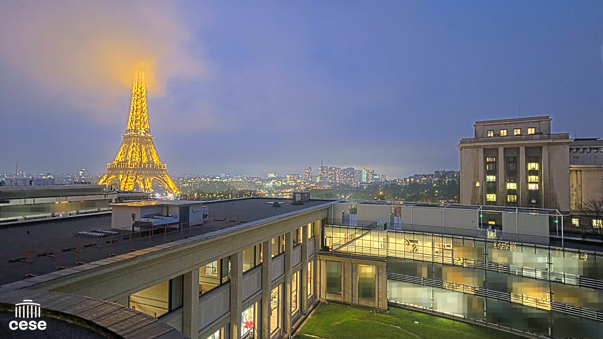 A nighttime view of the Eiffel Tower partially obscured by fog, with city lights and buildings in the foreground and background, under a twilight sky.
