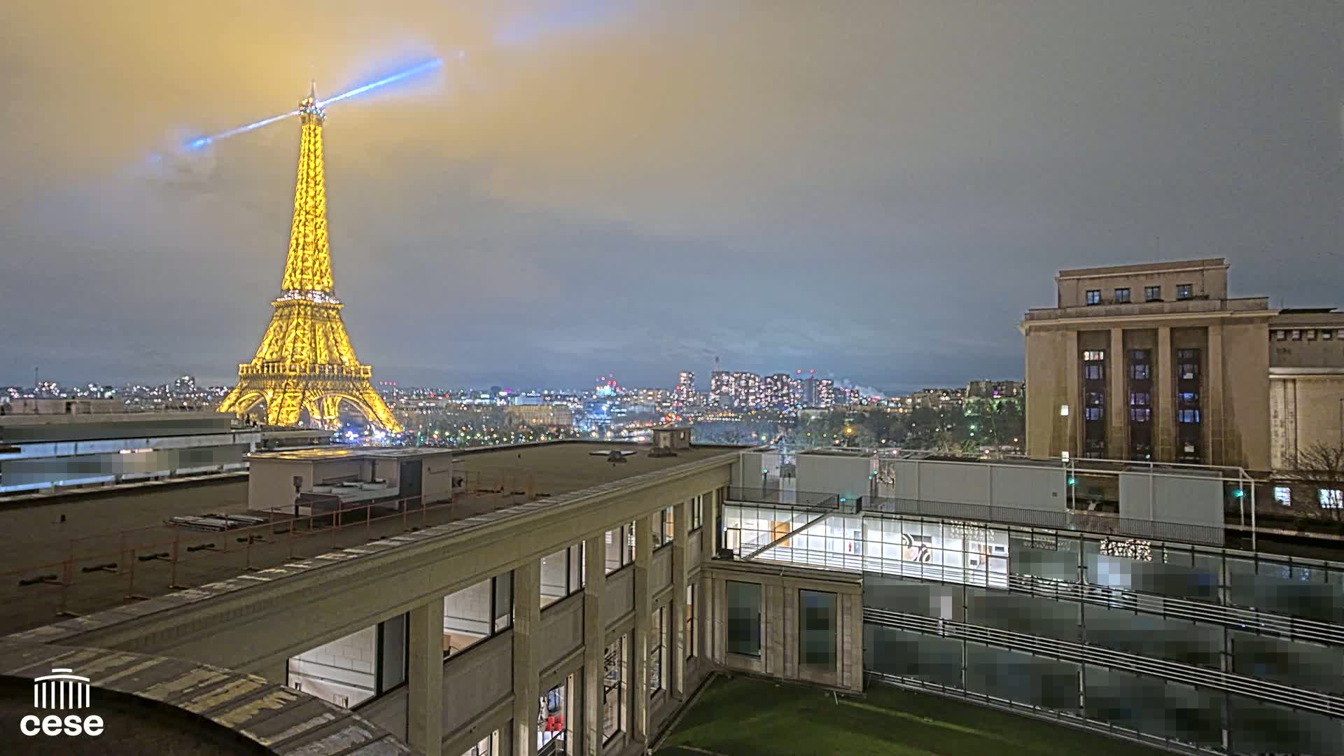A nighttime view of the Eiffel Tower illuminated, a city skyline, and a large building, under a cloudy sky.