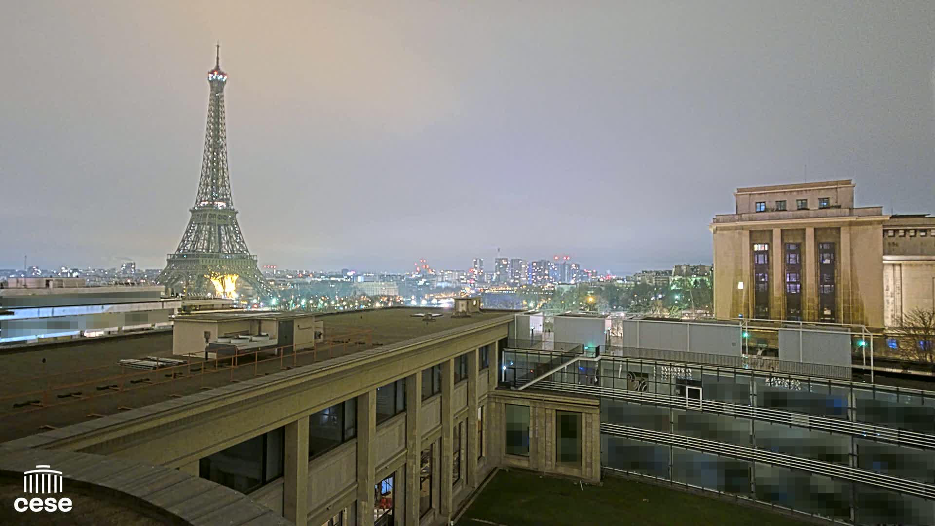 A nighttime view of the Eiffel Tower from a rooftop, overlooking a city under a hazy sky.