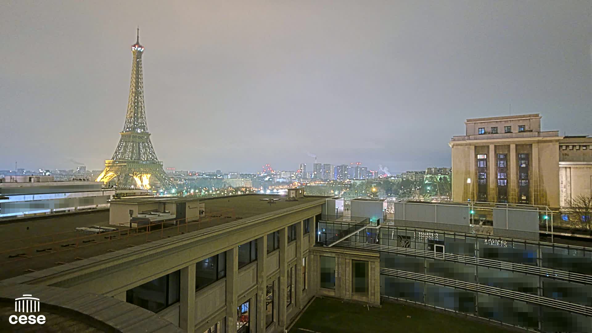 A nighttime view of the Eiffel Tower and a cityscape, under hazy or foggy conditions, from a rooftop vantage point.