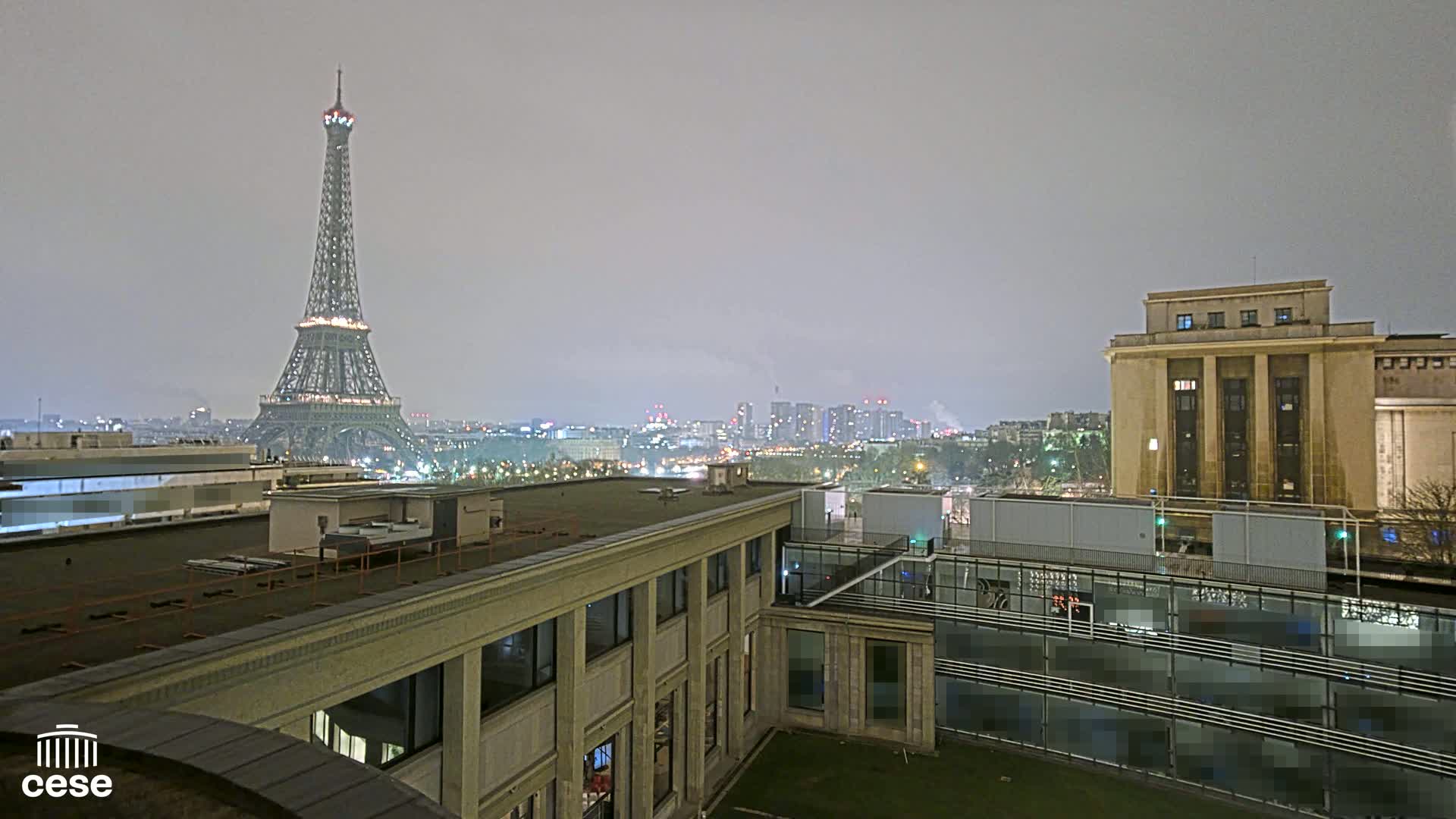 A nighttime view of the Eiffel Tower from a rooftop, under a hazy sky, with city lights and buildings in the distance.