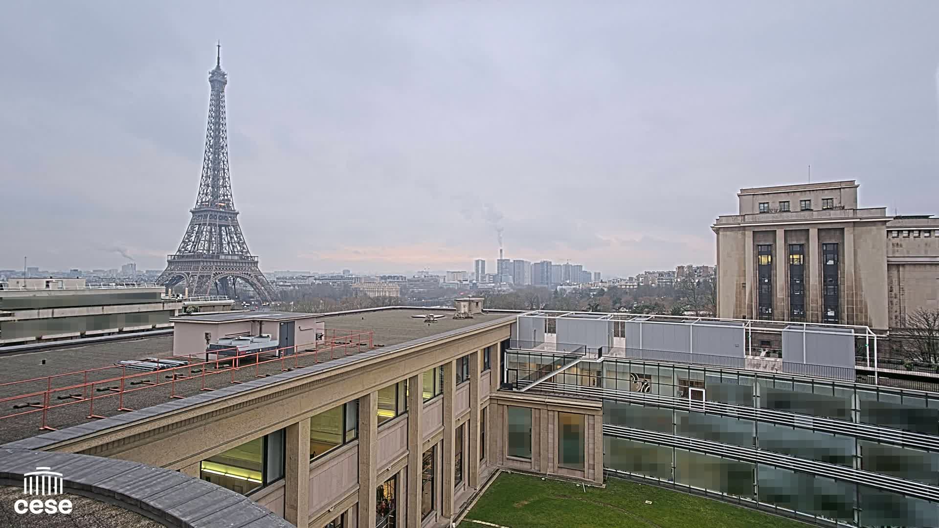 A view from a rooftop shows the Eiffel Tower in the distance on an overcast day, with several buildings and a small lawn in the foreground.