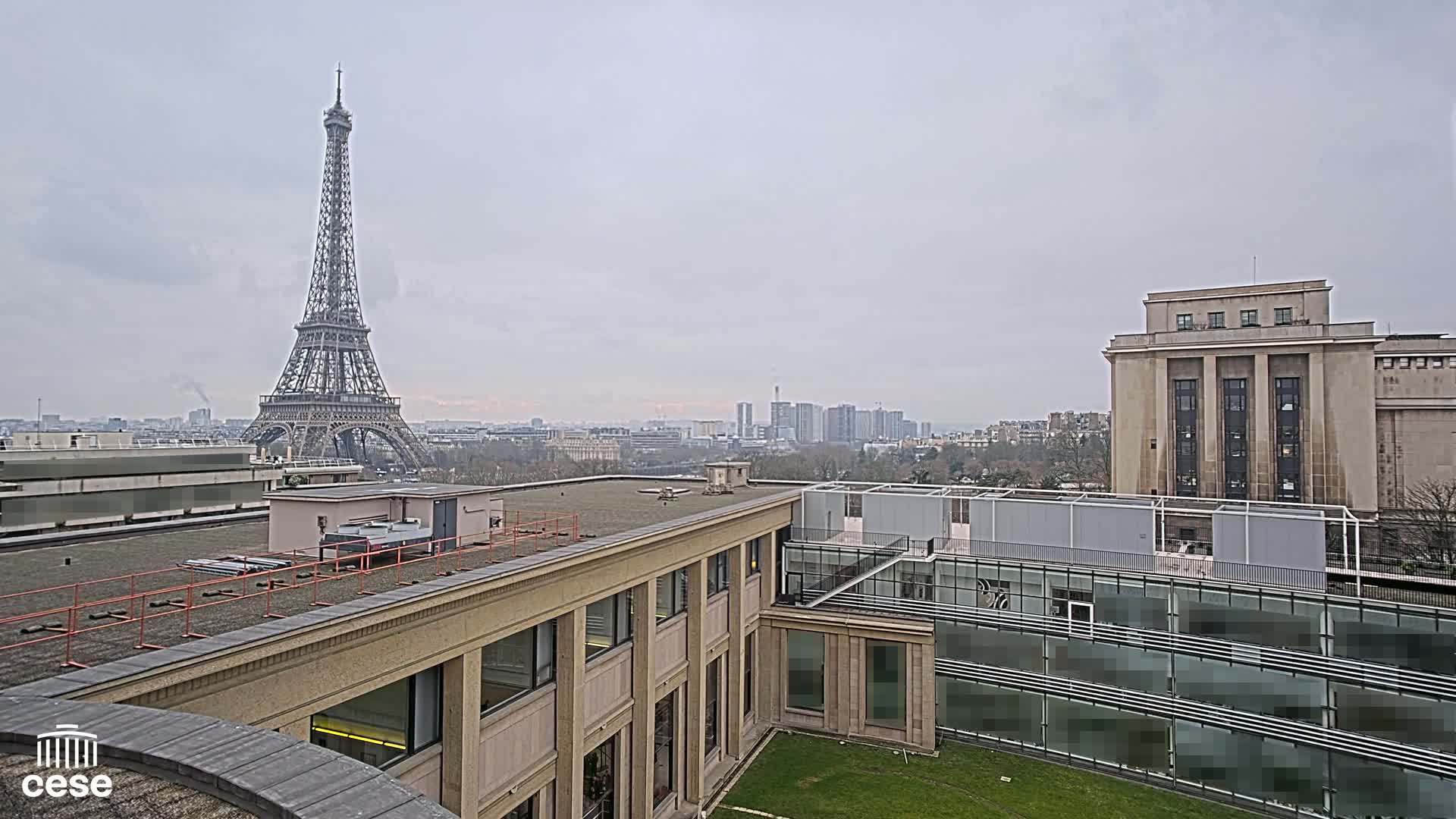 A view from a rooftop shows the Eiffel Tower in the distance under an overcast sky, with modern and classical buildings in the foreground.