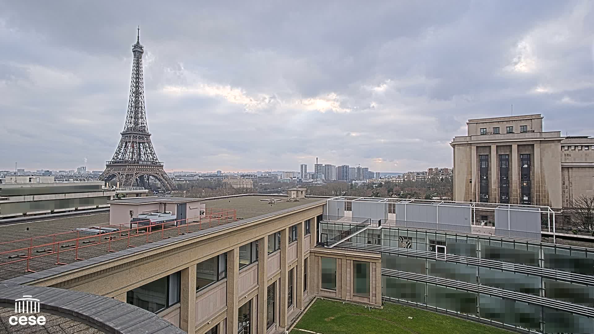 A view from a rooftop shows the Eiffel Tower in the distance under a cloudy sky, with other buildings and a small grassy courtyard in the foreground.