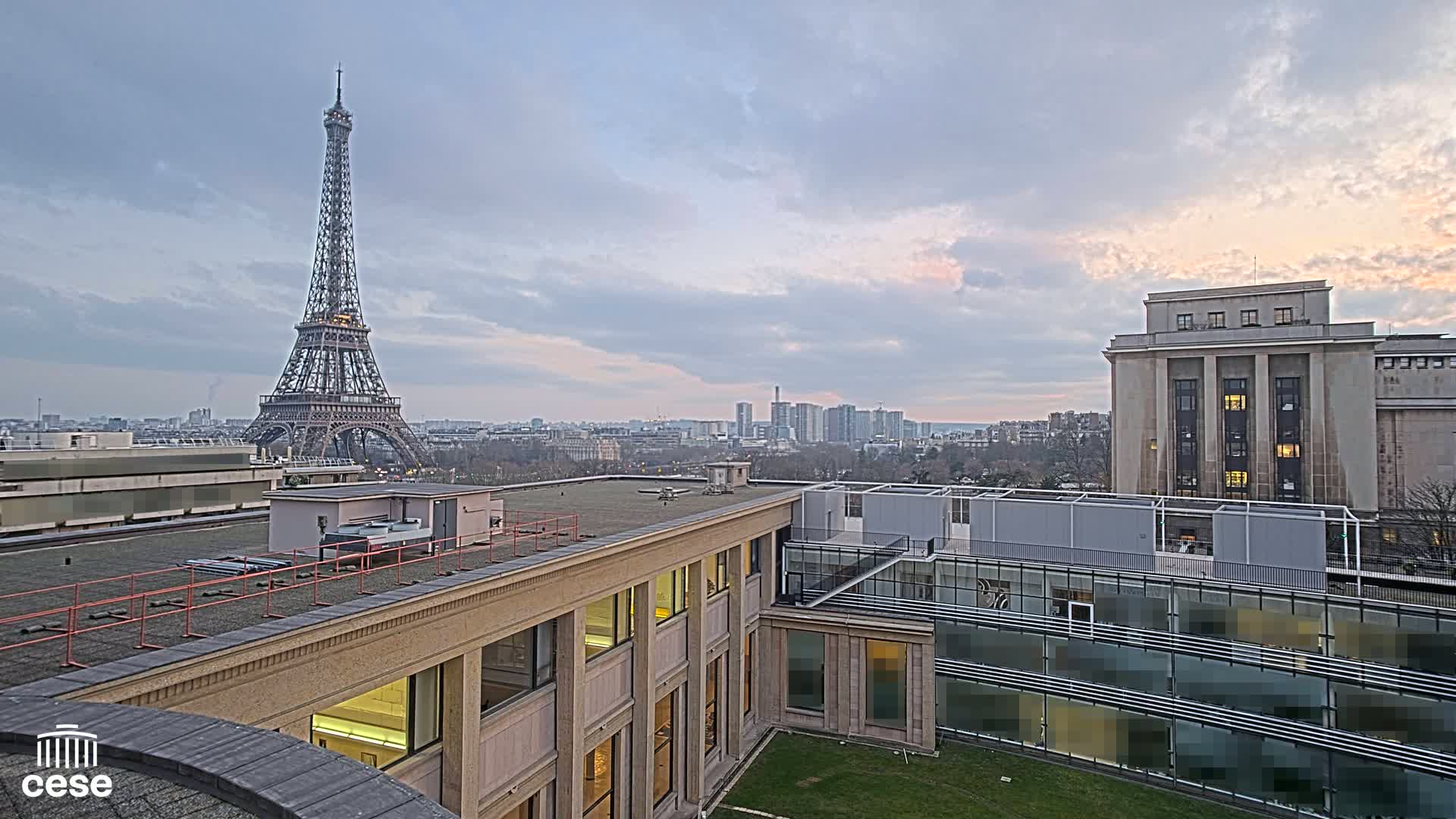 A rooftop view of the Eiffel Tower in the distance under a partly cloudy sky, overlooking a city skyline and other buildings.