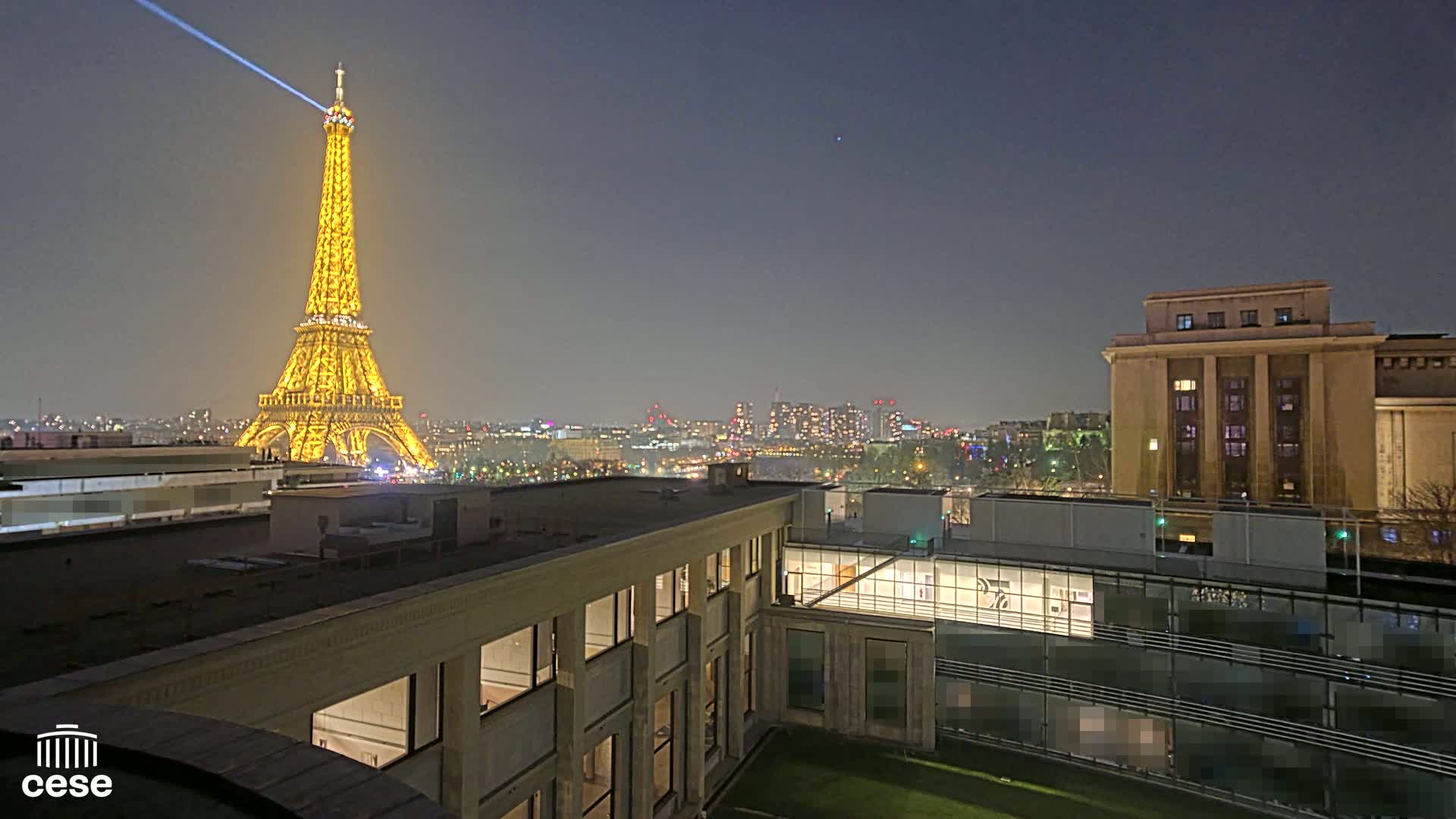 A brightly lit Eiffel Tower is visible at night from a rooftop vantage point overlooking a city skyline and a modern building.