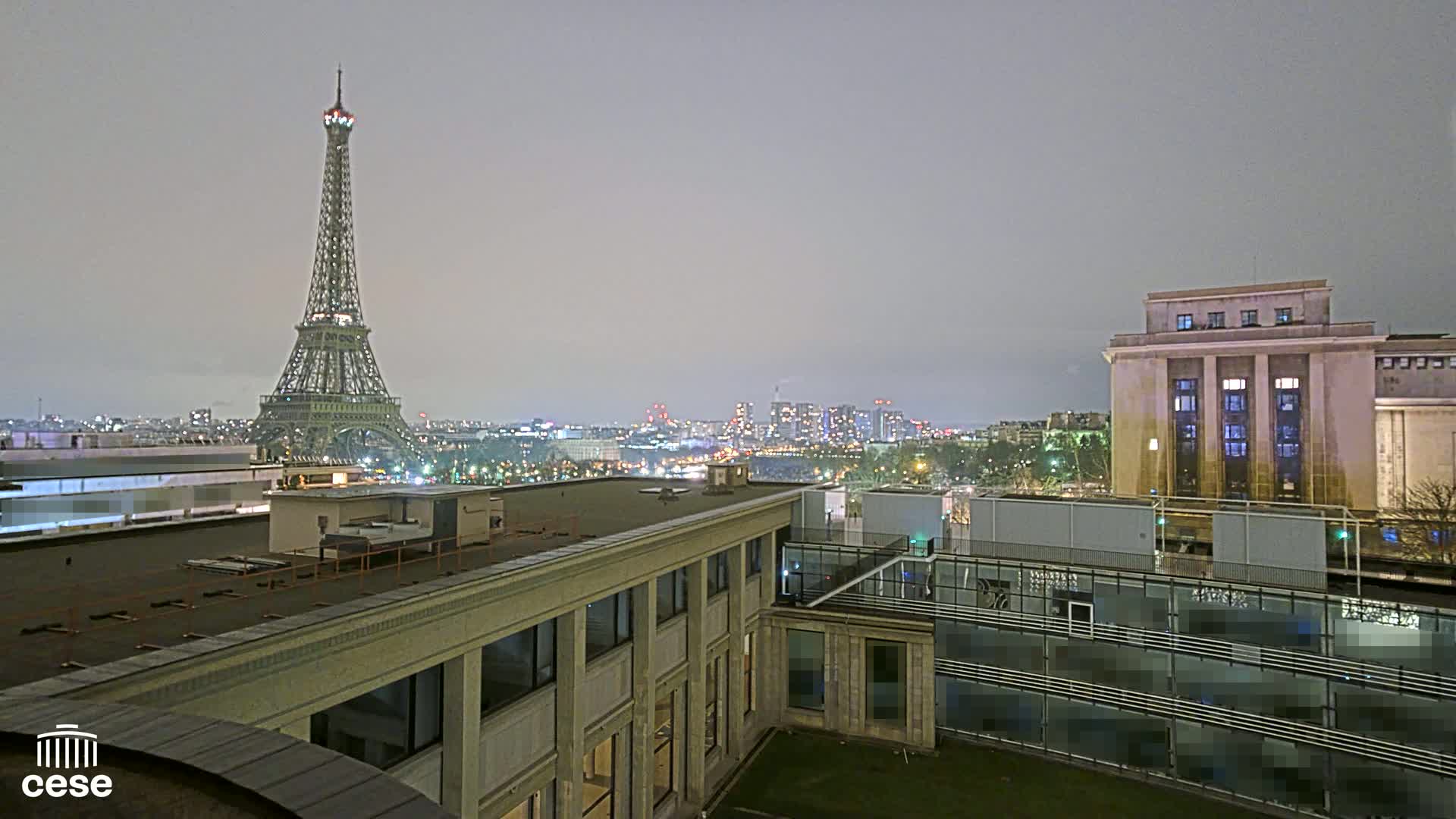 A nighttime view of the Eiffel Tower from a rooftop, with a city skyline visible in the distance under an overcast sky.