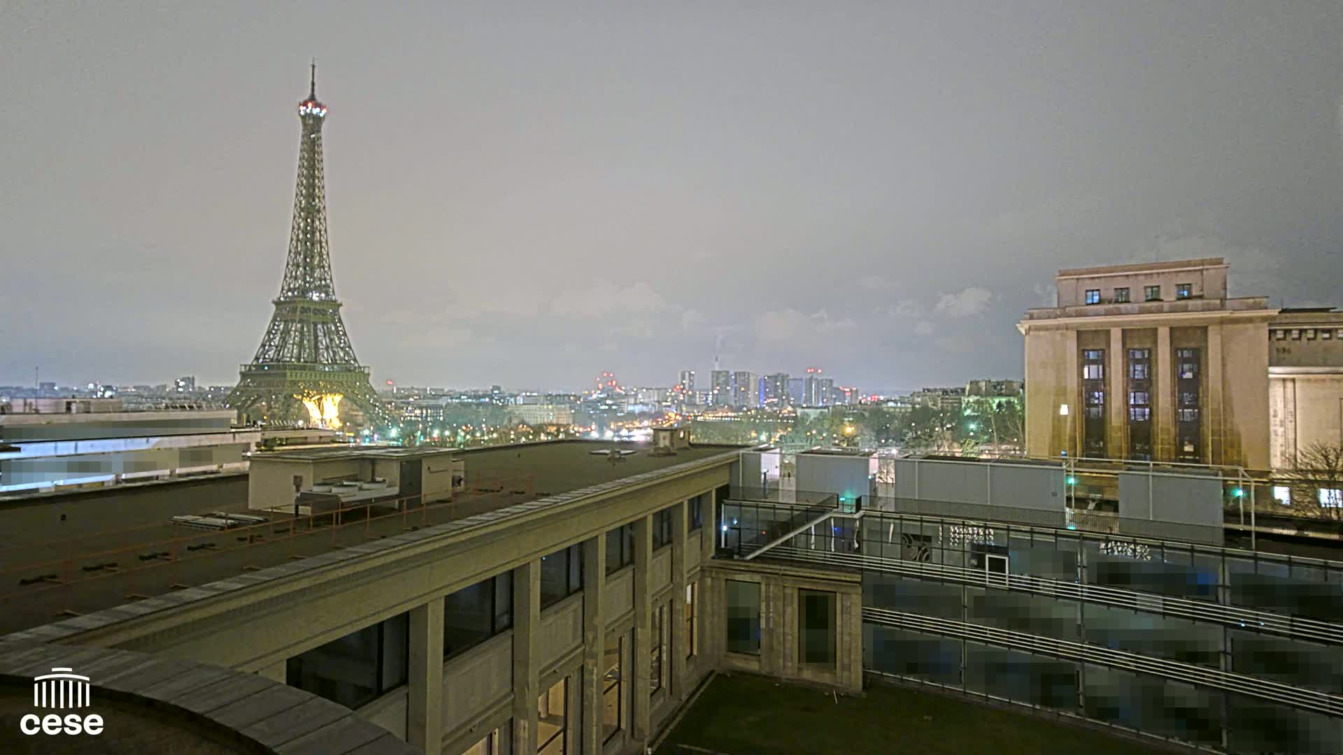 A nighttime cityscape view shows the Eiffel Tower in the distance under a cloudy sky, with city lights and buildings in the foreground.