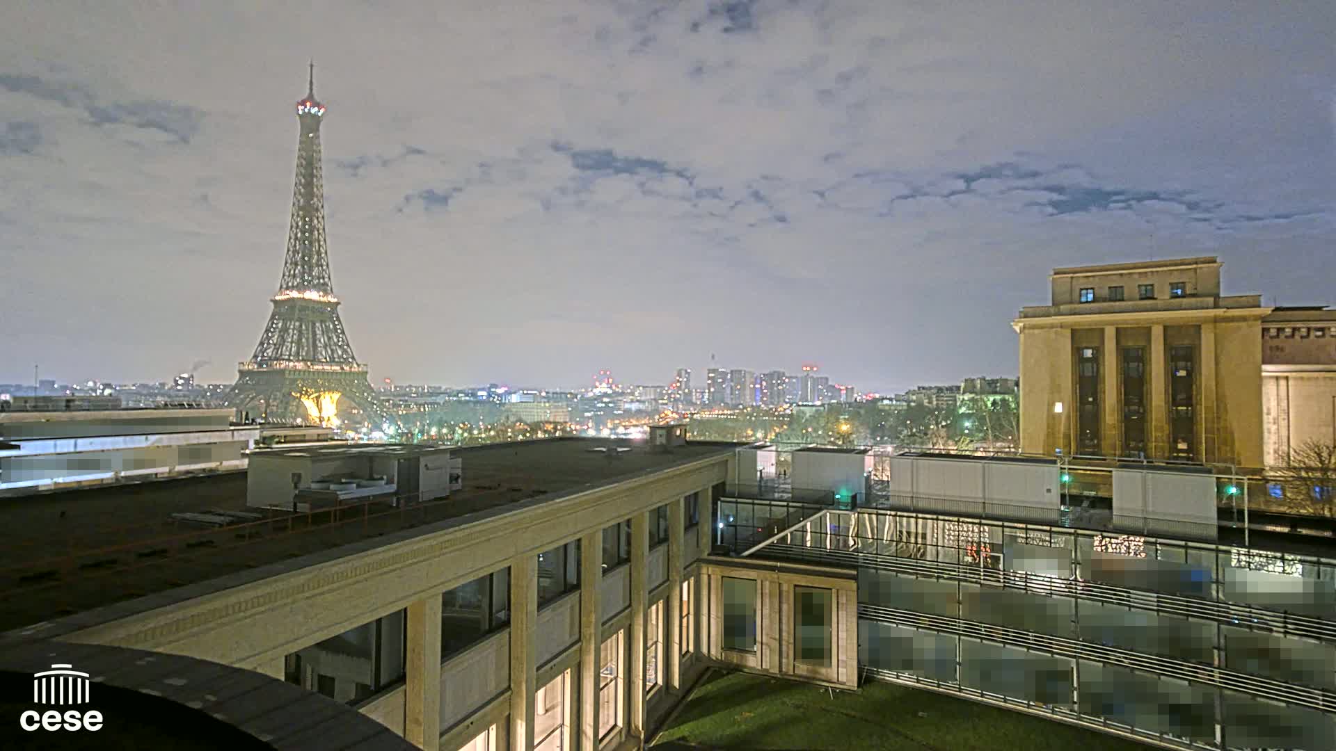 A nighttime view of the Eiffel Tower and Parisian cityscape from a rooftop, under a cloudy sky.
