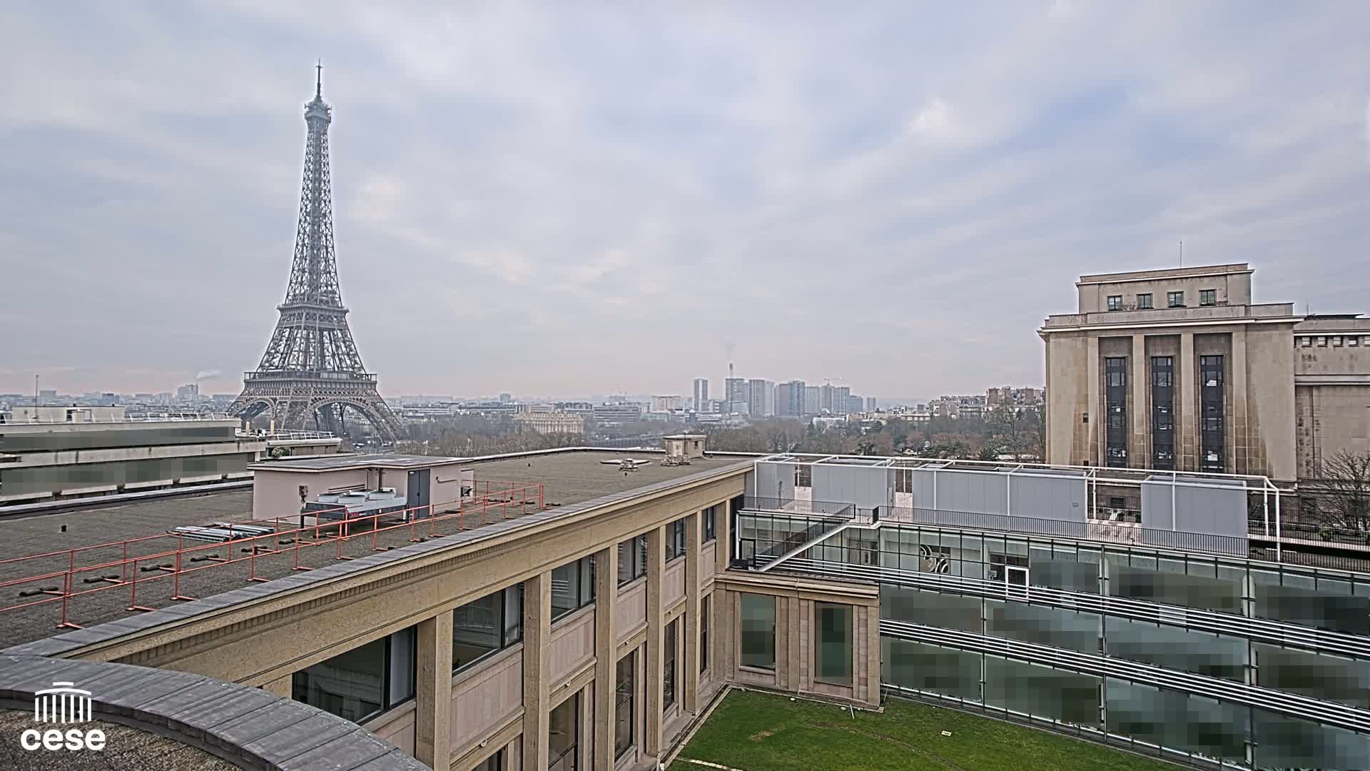 A view from a rooftop shows the Eiffel Tower in the distance under an overcast sky, with several buildings and a grassy courtyard in the foreground.